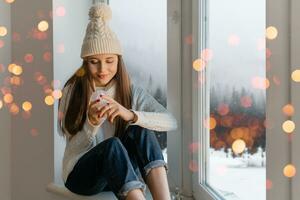 young attractive woman in stylish white knitted sweater, scarf and hat sitting at home on windowsill at Christmas holding glass snow ball present decoration, winter forest view, lights bokeh photo