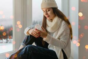 young attractive woman in stylish white knitted sweater, scarf and hat sitting at home on windowsill at Christmas holding glass snow ball present decoration, winter forest view, lights bokeh photo