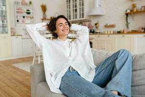 bonito joven mujer relajándose a hogar sentado en sofá, sonriente, feliz, gratis hora foto