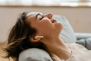 bonito joven mujer relajándose a hogar sentado en sofá, escuchando a música en auriculares foto