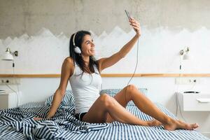 young attractive brunette woman sitting on bed in pajamas, listening to music photo