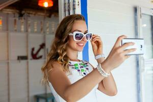 attractive woman in white dress in summer cafe in sunglasses using phone taking photo