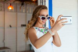 attractive woman in white dress in summer cafe in sunglasses using phone taking photo