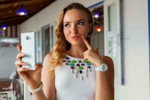 attractive woman in white dress in summer cafe in sunglasses using phone taking photo