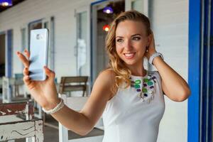 attractive woman in white dress in summer cafe in sunglasses using phone taking photo