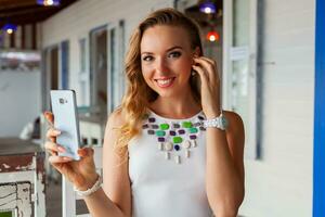 attractive woman in white dress in summer cafe in sunglasses using phone taking photo