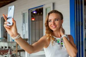 attractive woman in white dress in summer cafe in sunglasses using phone taking photo