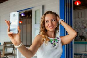 attractive woman in white dress in summer cafe in sunglasses using phone taking photo