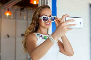 attractive woman in white dress in summer cafe in sunglasses using phone taking photo