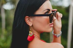 joven elegante sexy mujer en rojo verano vestir en pie en terraza en tropical hotel, palma arboles fondo, largo negro cabello, Gafas de sol, étnico pendientes, Gafas de sol, mirando adelante, cerca arriba foto