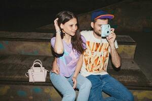 young stylish hipster couple in love, swag outfit, jeans, embrace, cool accessories, sitting happy, having fun, photo camera