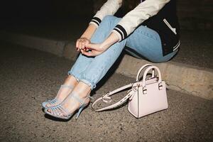 young stylish hipster woman, swag outfit, jeans, cool accessories, sitting on ground, pink purse, legs close up, details, shoes photo