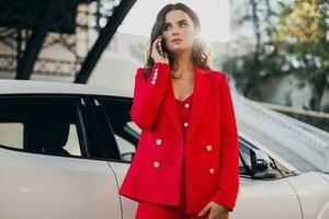 beautiful sexy woman in red suit posing at car talking on business on phone photo