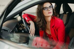 beautiful sexy rich business woman in red suit sitting in white car, wearing glasses photo