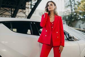 beautiful sexy woman in red suit posing at car talking on business on phone photo