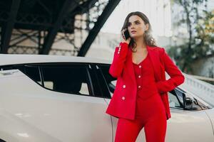 beautiful sexy woman in red suit posing at car talking on business on phone photo