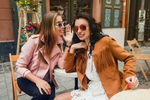 dos joven elegante mujer sentado a cafetería, hablando, chismoso, elegante de moda atuendo foto