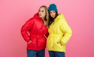 two attractive stylish women posing on pink background in colorful winter down jacket of red and yellow color, warm clothes fashion trend photo