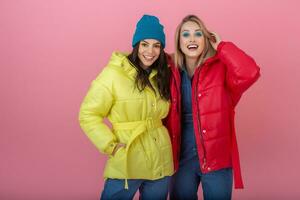 two attractive girl friends active women posing on pink background in colorful winter down jacket of bright red and yellow color having fun together, warm coat fashion trend, sincerely laughing photo