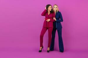 high fashion style two smiling attractive women on violet background in stylish colorful evening suits of purple and blue color, friends having fun together, fashion trend photo