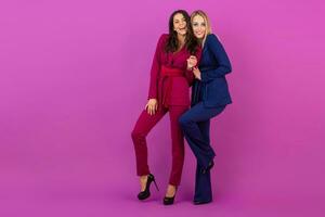 high fashion style two smiling attractive women on violet background in stylish colorful evening suits of purple and blue color, friends having fun together, fashion trend photo