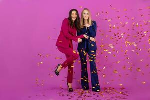 two attractive women celebrating new year on violet background in stylish colorful evening suits of purple and blue color, friends having fun together, fashion trend, golden confetti party mood photo