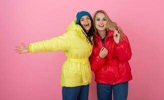 two excited attractive girl friends active women posing on pink background in colorful winter down jacket of bright red and yellow color having fun together, warm coat fashion trend photo