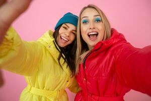 two attractive girl friends women taking selfie photo on pink background in colorful winter jacket of bright red and yellow color having fun together, warm coat sportswear fashion trend, crazy funny