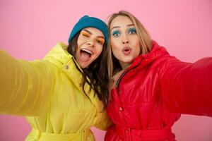 two attractive girl friends women taking selfie photo on pink background in colorful winter jacket of bright red and yellow color having fun together, warm coat sportswear fashion trend, crazy funny