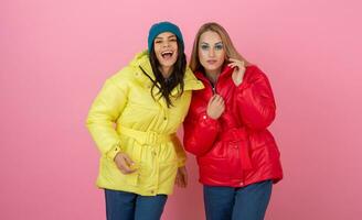 two excited attractive girl friends active women posing on pink background in colorful winter down jacket of bright red and yellow color having fun together, warm coat fashion trend photo