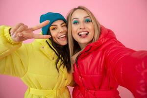 two attractive women posing on pink background in colorful winter down jacket of bright red and yellow color, friends having fun together, warm clothes fashion trend, crazy funny faces making selfie photo