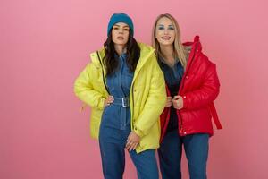 two excited attractive girl friends active women posing on pink background in colorful winter down jacket of bright red and yellow color having fun together, warm coat fashion trend photo