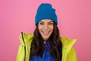 portrait of attractive smiling stylish woman posing in winter fashion look on pink background in bright neon yellow jacket, wearing blue knitted hat, dressed in warm clothes photo