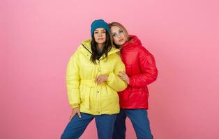 two attractive stylish women posing on pink background in colorful winter down jacket of red and yellow color, warm clothes fashion trend photo