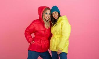 two excited attractive girl friends active women posing on pink background in colorful winter down jacket of bright red and yellow color having fun together, warm coat fashion trend photo