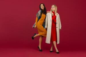 two stylish women in autumn winter fashion dress and coat posing isolated full heigth on red studio background photo