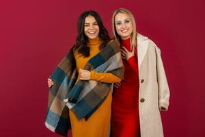 two smiling attractive stylish women in autumn winter fashion dress and coat posing isolated on red studio background photo