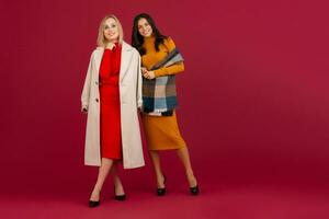 two stylish women in autumn winter fashion dress and coat posing isolated full heigth on red studio background photo