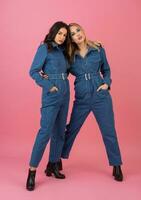 two excited attractive girl friends active women posing on pink background in denim overall having fun together fashion trend photo