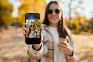 atractivo joven mujer caminando en otoño vistiendo chaqueta utilizando teléfono foto