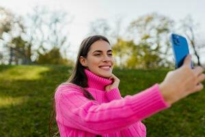 joven sonriente mujer en rosado suéter caminando en verde parque utilizando teléfono foto