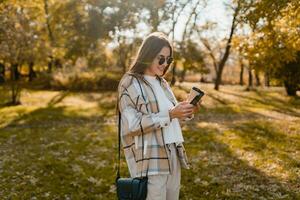 atractivo joven mujer caminando en otoño vistiendo chaqueta utilizando teléfono foto