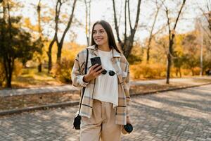 atractivo joven mujer caminando en otoño vistiendo chaqueta utilizando teléfono foto