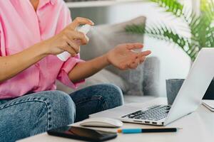 close up hands of woman spray sanitizer antiseptic at workplace at home working online on laptop photo