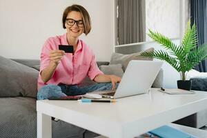 woman in pink shirt sitting relaxed on sofa at home at table working online on laptop photo