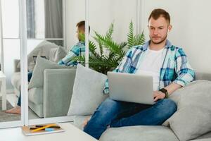 handsome smiling man in shirt sitting relaxed on sofa at home at table working online on laptop photo