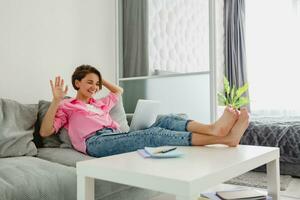 attractive smiling woman in pink shirt sitting relaxed on sofa at home at table working online photo