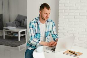 man in shirt sitting in kitchen at home at table working online on laptop photo