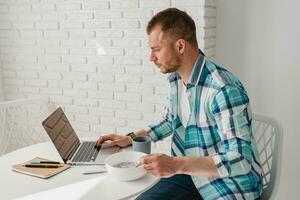 man in shirt sitting in kitchen at home at table working online on laptop photo