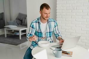 handsome smiling man in shirt sitting in kitchen at home at table working online on laptop photo